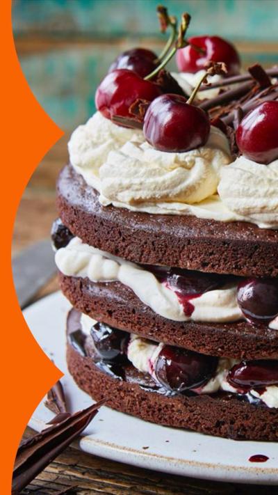 A Black Forest gâteau cake - with three layers of chocolate cake, cream and cherries - sits on a white plate on a wooden table 