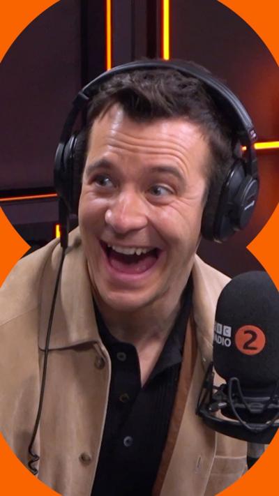 Luke Thompson has a big smile. He has short brown hair and wears a camel-coloured jacket and black headphones.