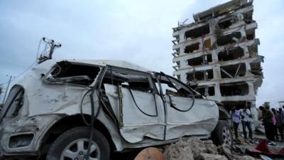 Somali government soldiers and journalists stand near a car destroyed in front of the Jazeera hotel after an attack in Somalia's capital Mogadishu