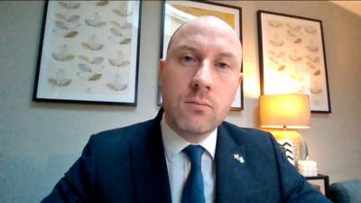 Scottish Health Secretary Neil Gray wears a navy blue suit and sits in front of a gray wall with three framed floral prints.