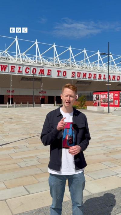 Andy Watson, a BBC reporter standing in an open paved plaza outside a football stadium, facing the camera and gesturing with one hand. Behind him, large red letters spell out “WELCOME TO SUNDERLAND” across the stadium front. The stadium features white metal framework along the roofline and numbered entrances below. To the right, a sign advertises a “Fan Zone,” and tall lampposts line the area under a clear blue sky.