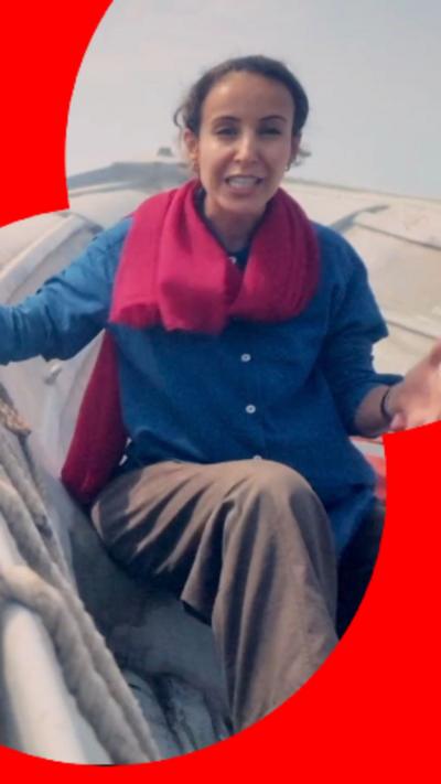 Nawal wears a scarf, shirt and trousers as she sits in a boat in the strait with a container ship seen in the background.