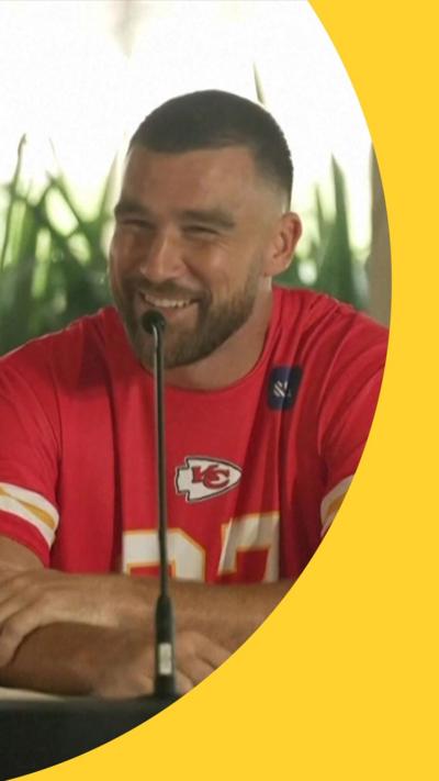 Travis Kelce behind a microphone wearing a Kansas City Chiefs shirt