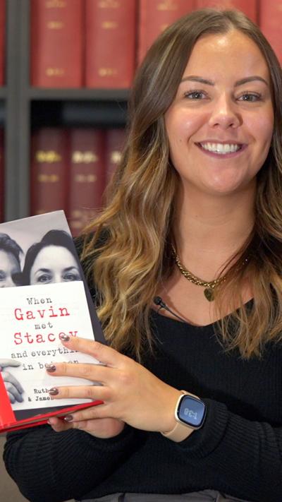 Tink, a BBC journalist with blonde and brown hair. She smiles at the camera and has  a black top on. She holds a book to the camera, the title of the book is When Gavin Met Stacey.