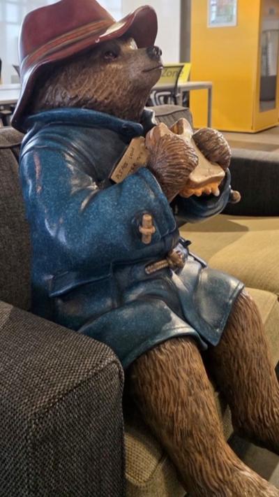A paddington bear statue sits on a grey chair. The statue wears the bear's signature red hat and blue coat and holds a marmalade sandwich with a bite taken out of it.