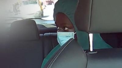 Axel Rudakubana, wearing a green hoodie and a blue mask across his mouth and nose, sits in a taxi with grey seats