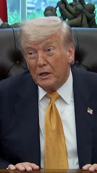 Donald Trump wears a yellow tie with a white shirt and blue suit while sitting behind his desk in the Oval Office.