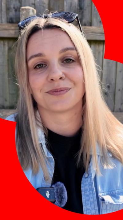 A woman with long blonde hair smiles into the camera. She is wearing a black T-shirt under a light blue denim overshirt.