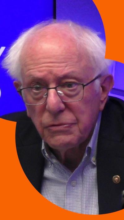 Bernie Sanders wears a dark suit jacket and a light checked shirt; he has white hair and black rimmed glasses