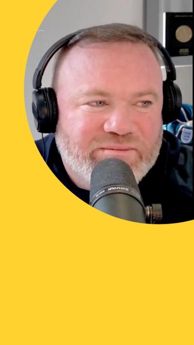 Wayne Rooney, wearing headphones, speaks into a mic. He has short brown hair and a grey beard 