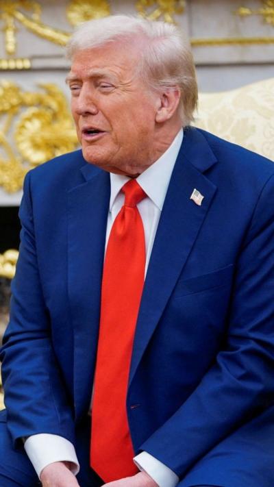 Donald Trump sits in the Oval Office wearing a blue suit and red tie as he speaks towards the press (not pictured).