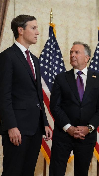 Jared Kushner and Steve Witkoff in black suits, stood next to two US flags