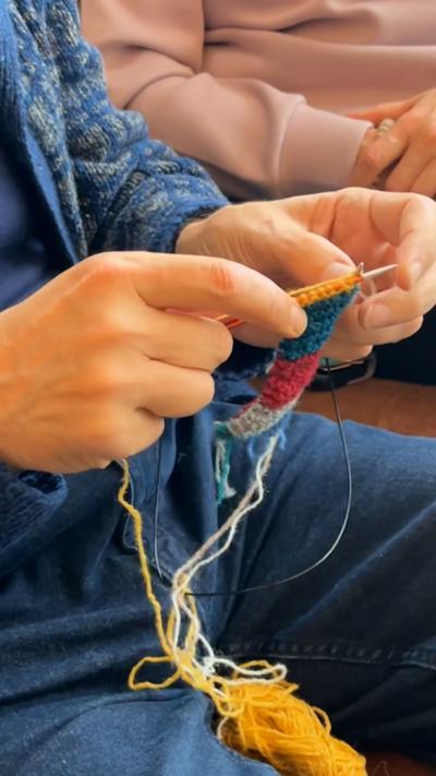 Man's hands knitting segments of a yellow, blue, red and grey wool hat