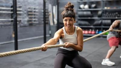 A woman smiles, with her hair in messy top knot, playing tug of war at a cross training gym.