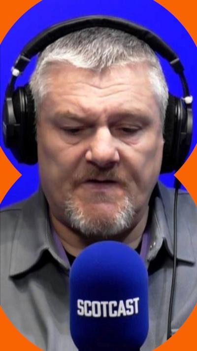John Davidson sits in front of a radio studio microphone, he wears headphones and a grey shirt