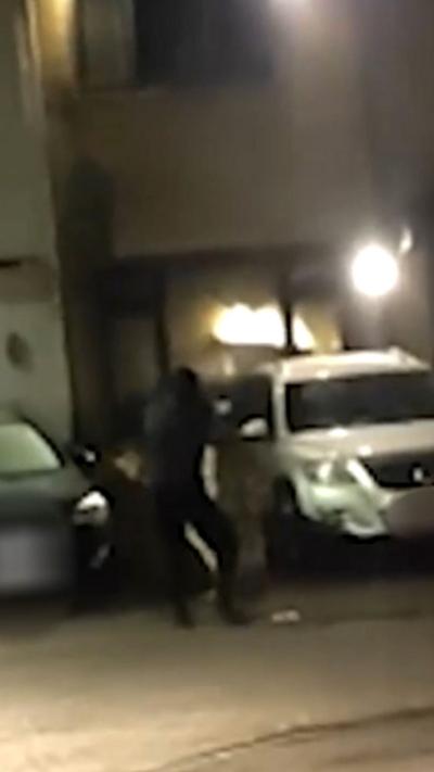 A man dressed in black firing a gun in a dark street in front of a house with a silver SUV.