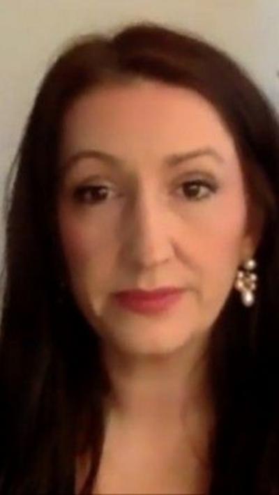 The image is a screenshot from a video call with Emma Little-Pengelly. She has long brown hair, brown eyes, and is wearing long pearl earring.