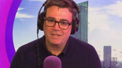 Andy Burnham in a dark shirt and glasses, sat at a Radio Manchester microphone