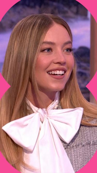 Sydney Sweeney smiles in The One Show studio, wearing a large white bowtie and a grey cardigan. She has long brown hair