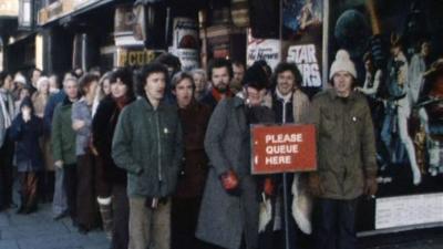 A queue of people, all wearing warm coats, standing in front of Star Wars posters outside a cinema.