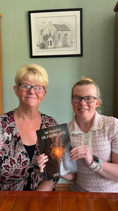 A mother and daughter, both wearing glaseses, sat down with a book called Between Heartbeats. The mother has blonde hair and a purple, grey and black patterned top. Her daughter is wearing a pink and white short-sleeved top.