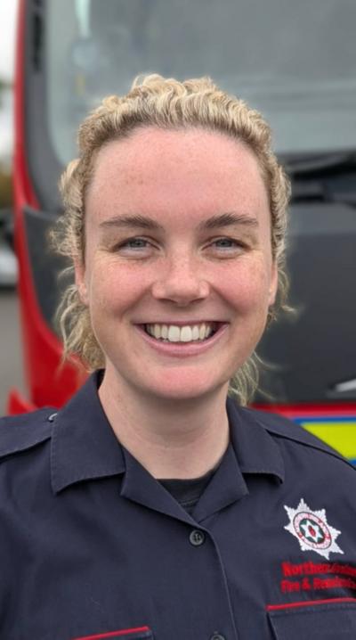 Ciara McGurk has curly blonde hair and is wearing a NIFRS navy uniform. She is smiling and standing in front of a fire engine.