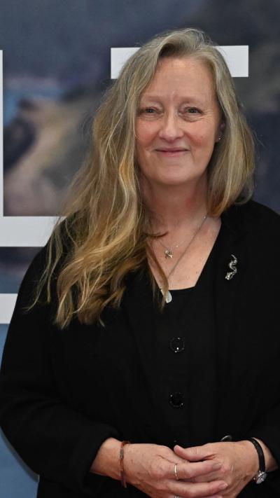 Salt Path author Raynor Winn pictured in front of a promotional book for her novel. She has blonde long hair which is swept onto her one shoulder and smiles at the camera.