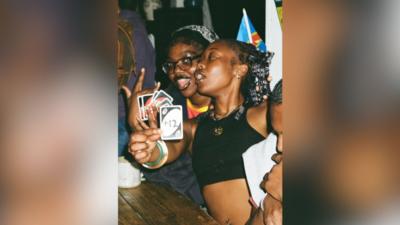 Two young women playing uno, one of them is showing a card with '+12' written on it