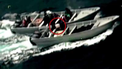 Two figures in black carry a large white bag circled in red between two motorboats from the point of view of a camera scope.