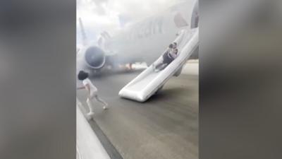 A man slides down an inflatable evacuation slide from a plane as a carrying a child. Another child runs away from the slide. There is smoke in the background, coming from the plane.