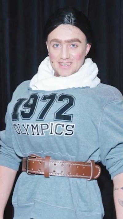 Roman Kemp dressed as Miss Trunchbull from Matilda. He wears a black wig and a 1972 Olympics sweater with a big brown belt