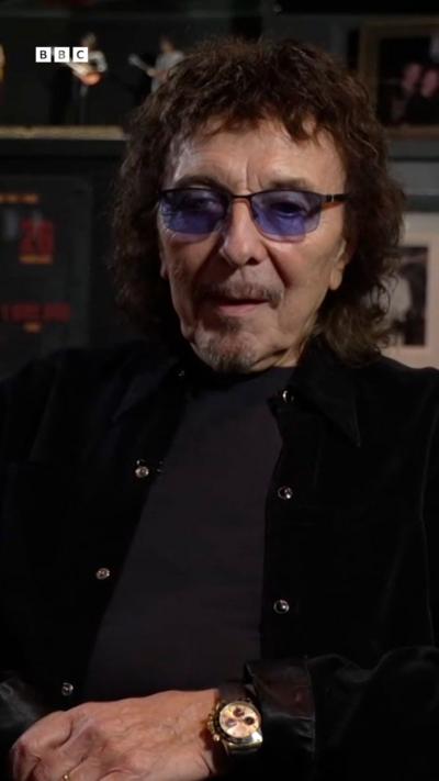 A with dark brown hair, shaded glasses, black jacket and black top, sat on a sofa in a dark room. A guitar is on a stand next to him and there are various ornaments and photos in the background