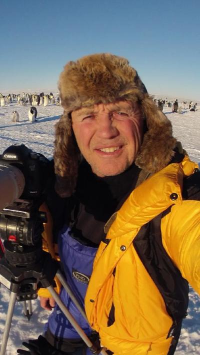 Doug Allen stood in an icy area, wearing a thick yellow coat and dearstalker hat. Behind him are penguins and he is holding a camera.