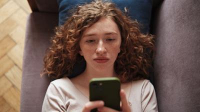 A woman lying on a sofa holding her phone up looking bored