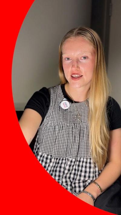 A young woman with long blonde hair, wearing a black and white checked sleeveless top over a black t-shirt. 