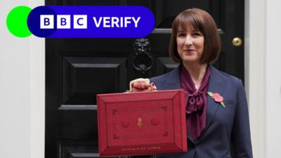 The Chancellor of the Exchequer Rachel Reeves stands in front of the door to 11 Downing Street. She is wearing a navy blue suit with a dark purple blouse, and is wearing a poppy. The photo is from the Autumn Budget announcement in 2024. She is holding up a red box to the press to symbolise the new budget of the UK government. The BBC Verify logo is in the top left corner.