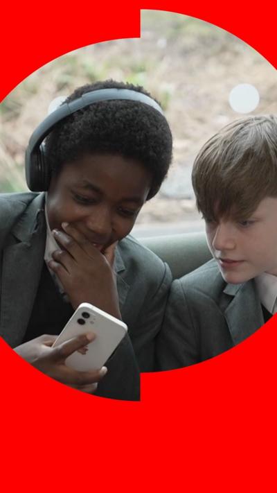 Two boys looking at a mobile phone. They are wearing grey school uniform. One boy is wearing headphones.