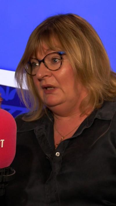 A woman speaking into a studio microphone during a broadcast, seated against a blue backdrop, wearing a dark shirt.