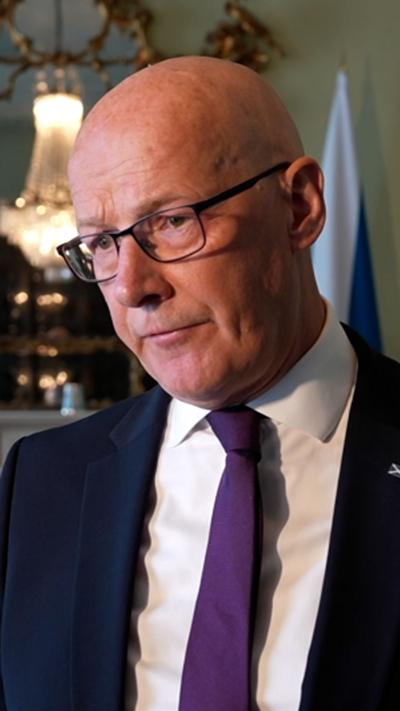 John Swinney wears glasses, a dark suit, white shirt and purple tie