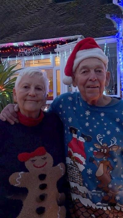An older couple wearing Christmas jumpers stand in front of their Christmas light covered bungalow.