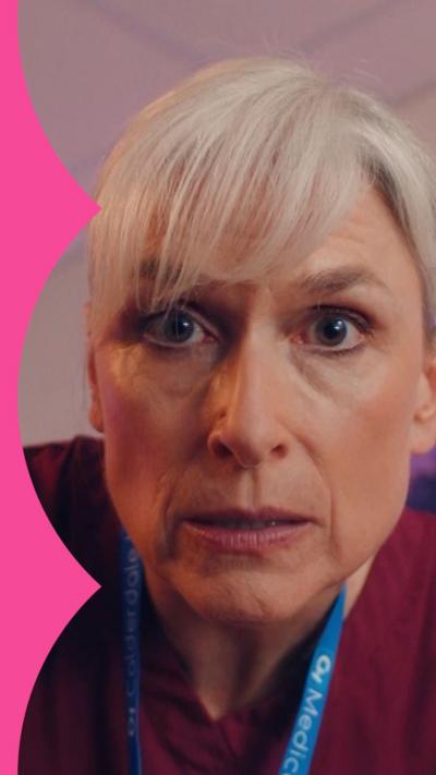 Close up of woman with a grey fringe and purple scrubs, who peers at the camera.