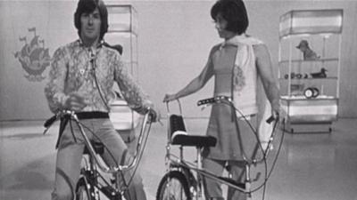 Black and white image of Peter Purves and Valerie Singleton on chopper bikes in the Blue Peter studio.  In the background are a couple pieces of scenary and a Blue Peter ship logo hanging from the ceiling.  Peter is astride his bike and looking directly down the lens.  Valerie is standing beside her bike and looking at Peter.