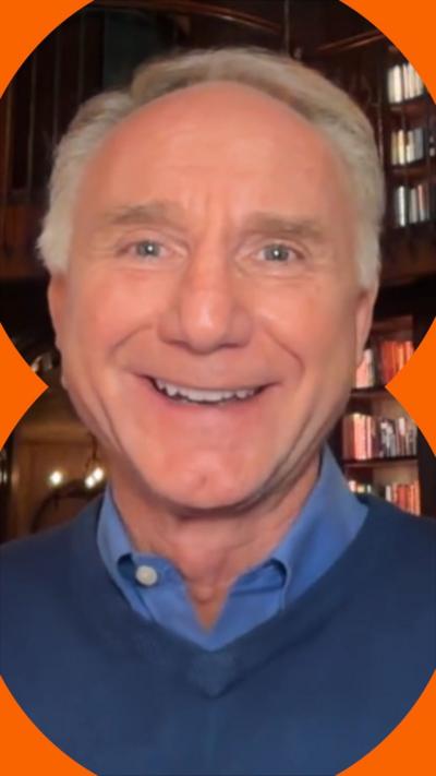 Dan Brown smiles. He has short grey hair and wears a blue shirt under a blue jumper. He is in a room full of bookshelves
