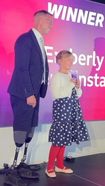 Aaron Phipps stands in front of a large sign which says 'winner'. Next to last year's Young Hero Award winner, Emberley. Aaron wears a blue suit with a white shirt and silver tie. He has short dark brown hair and wears glasses. Emberley is standing next to him holding up her award. She hair blonde hair tied back into a low bun, with a blue and white polka dot dress on and white cardigan, red tights and pink ballerina pumps.