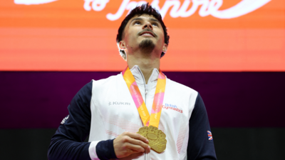 Jake Jarman is standing in the middle of the image looking up. He has short black hair and a beard and is wearing a white and navy tracksuit top. He has a gold medal around his neck.