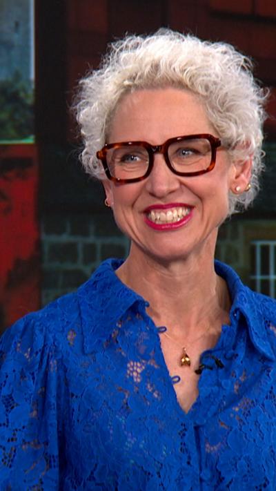 Anna Campbell-Jones is in a BBC news studio, wearing glasses and a blue shirt, she has grey hair and is smiling