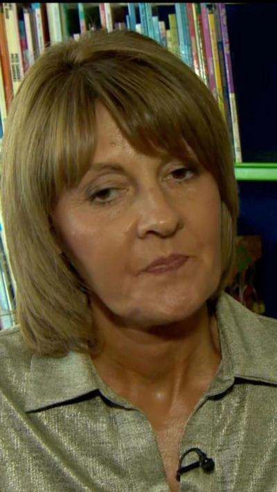 Jenny Bowles has chin-length light brown hair with a fringe. She is wearing a beige top with a collar. She looks emotional. 