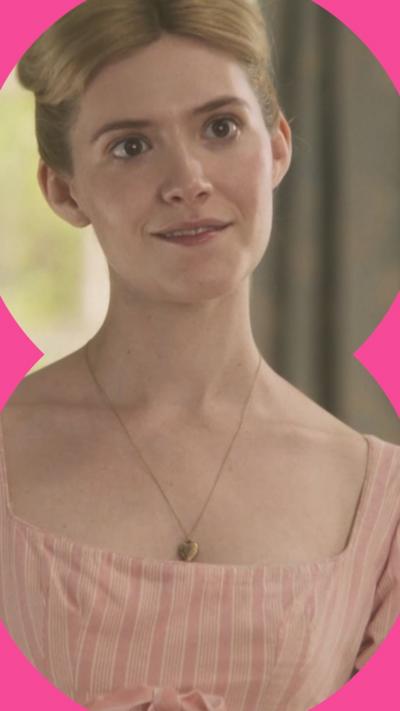 A woman with blonde hair smiles at someone off camera, she wears a pink dress