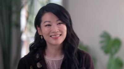 Headshot of Arden Cho smiling to the side of the camera. She has long, wavy, black hair and is wearing several gold, jewelled earrings. She is wearing a sheer cream top with brown embroidery and a brown jacket with gold brooches. She is pictured in a room with cream walls and window behind her, with large cheese plant.