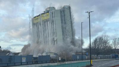 The grey tower block is being demolished in a plume of smoke. 